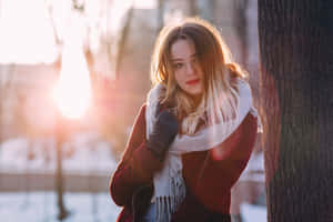 身穿冬季外套和圍巾的女性靠在雪地公園的樹上，陽光照射。 — 酷寒冬季 壁紙