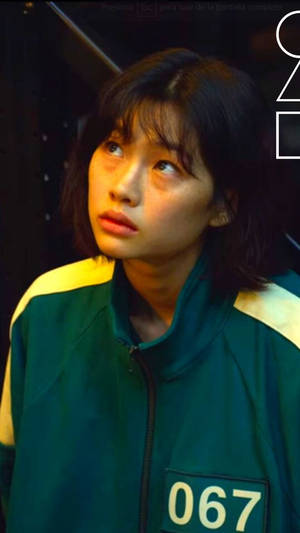 身穿綠色和黃色外套、號碼067的年輕女性向上看，表情憂慮。 — 067 魷魚遊戲 壁紙
