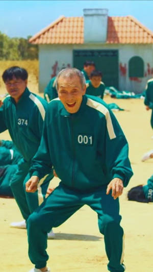 一位身穿綠色號碼運動服的長者在遊戲節目場景中與其他參賽者一起大笑。 — 067 魷魚遊戲 壁紙