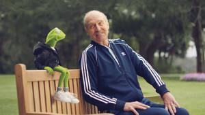 一位身穿藍色運動服的長者與穿著連帽衫的青蛙柯米特坐在長椅上。 — 運動 壁紙