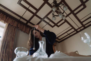 身穿藍色長袍的女性在室內手持鐘擺懸於擺放玻璃器皿的桌子上方。