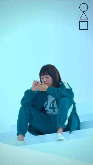 年輕女性身穿青綠色運動服，坐在極簡風格工作室的白色台階上，周圍有幾何形狀。 — 067 魷魚遊戲 壁紙