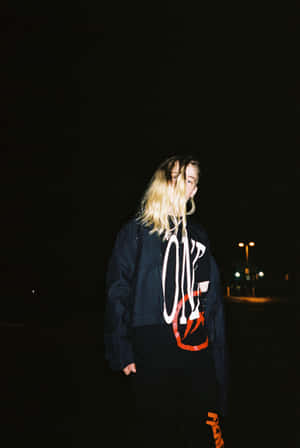 金髮人士在夜晚的城市環境中穿著圖案連帽衫和牛仔外套。 — Vlone 壁紙