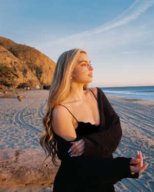穿黑色服裝的女性站在日落沙灘上，背後有海洋和山丘 — 艾迪生·雷 壁紙