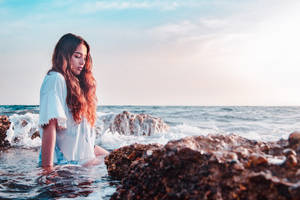 年輕女子在日落時坐在海邊岩石附近的波浪中 — 日落 壁紙
