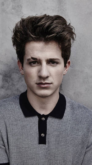 穿著灰色和黑色Polo衫的年輕男子在灰色牆壁前直視鏡頭。 — 查理·普斯 壁紙