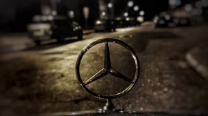 Mercedes-Benz 標誌在濕漉漉的人行道上，夜晚，後面有模糊的汽車。 — Mercedes Benz C300 壁紙
