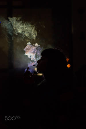 在昏暗的房間裡，一個人呼出彩色煙霧，周圍有發光的燈光。 — 抽菸 壁紙