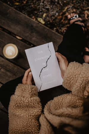 一個人在秋天戶外喝咖啡時閱讀名為《The Strength in Our Scars》的書。 — 書本美學 壁紙