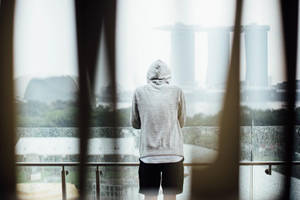 穿著連帽衫的人從雨中的陽台透過模糊的垂直欄杆凝視新加坡天際線。 — 孤獨悲傷 壁紙