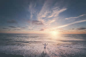 日落時分，一個人站在海浪中高舉雙臂 — 海灘桌面 壁紙