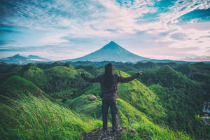 一個人在長滿草的山丘上張開雙臂，俯瞰日出時的火山。 — 山坡 壁紙