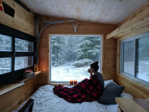 小屋裡的人一邊喝茶一邊透過窗戶觀看白雪覆蓋的森林 — 冬季風景 壁紙