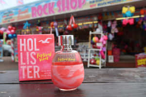 粉紅色Hollister香水瓶和盒子放在色彩繽紛商店外的木桌上。 — 3:2 壁紙