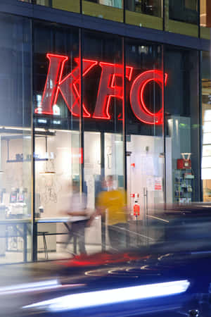 一個發光的紅色KFC標誌在玻璃外牆的餐廳中，外面有模糊的人影和汽車移動。 — 榮耀MagicPad 3 壁紙
