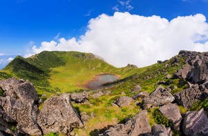 一個寧靜的火山口湖，被綠色山丘和灰色岩石環繞，在清澈的藍天下。 — 제주도 壁紙