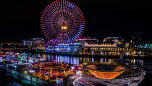 夜晚燈火通明的遊樂園，巨大的摩天輪和色彩繽紛的遊樂設施倒映在河面上。 — 霓虹燈 壁紙