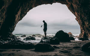 人站在海蝕洞內，望著海洋，附近有一隻鳥飛過。 — 海灘 壁紙