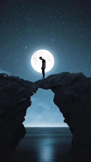 夜晚，一個人站在岩石拱門上，滿月高懸於海面上空的剪影。 — 月光 壁紙