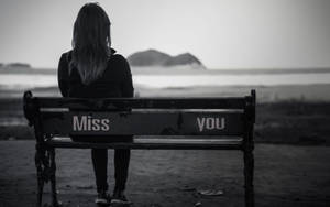 一個人獨自坐在海邊的長椅上，上面寫著「Miss you」。 — 心碎 壁紙