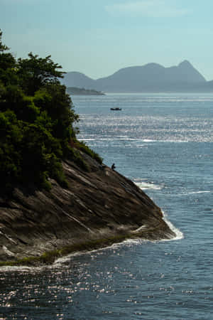 一個人坐在岩石懸崖邊緣，俯瞰海洋，遠處有山脈。 — 海洋 壁紙