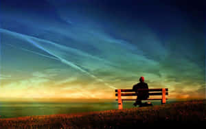 戴紅帽的人坐在長椅上，面向夕陽下的海洋的剪影。 — 獨自一人 壁紙