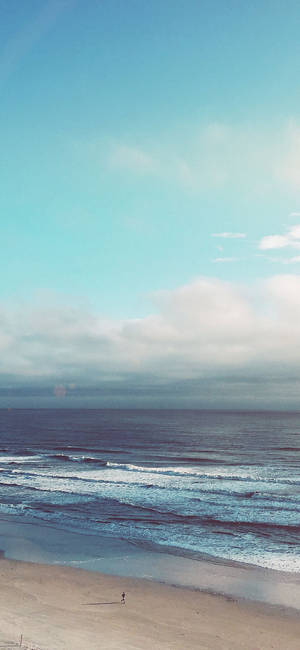 一個孤獨的人在海灘上行走，有海浪和多雲的天空。 — 海灘 iPhone 壁紙