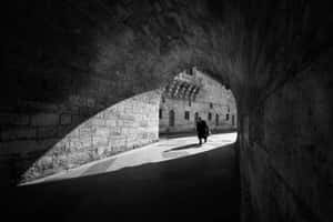 一個人穿過石頭隧道，強烈的陰影和光線顯露出盡頭的建築物。 — 民間藝術 壁紙