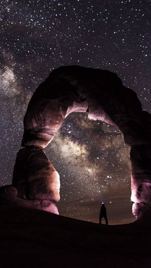 夜晚，一個人站在巨大岩石拱門下的剪影，可見星星和銀河。 — 自然 iPhone 壁紙