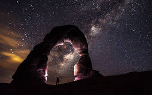 夜晚，一個人站在天然石拱門下，可見星星和銀河。 — 星空動畫 壁紙