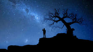 人站在懸崖上，星空下有銀河和光禿禿的樹 — 夜晚 壁紙