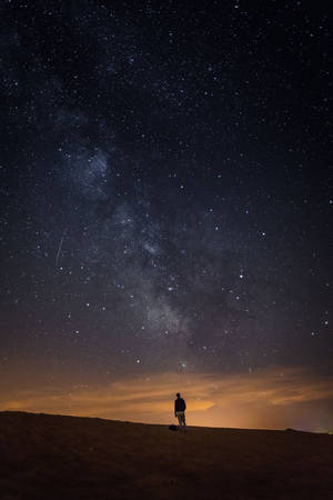 一個人的剪影站在沙漠沙丘上，在銀河和流星點綴的星空下。 — 暗色 壁紙