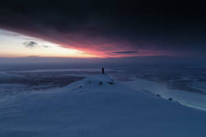 一個人獨自站在白雪覆蓋的山頂，在夕陽下俯瞰冰凍平原。 — 雪景 GIF 壁紙