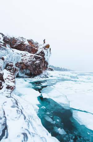 人站在積雪懸崖邊緣，俯瞰結冰的藍綠色湖泊。 — 冬季風景 壁紙