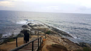 人物走下樓梯到岩石海邊，下方有海浪拍打。 — 水 壁紙