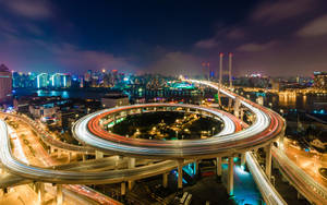 夜晚閃爍的高速公路交匯處，移動車輛的光跡和背景的城市天際線。 — 城市風景 壁紙