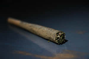 一個已點燃的大麻煙捲在深色反光表面上，煙霧裊裊升起。 — 大麻 壁紙