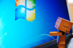帶有亞馬遜標誌的紙板Danbo在顯示Windows 7桌面的筆記型電腦旁邊微笑。 — 酷炫桌面 壁紙