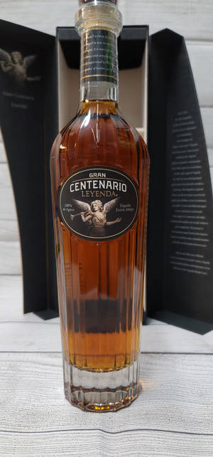 一瓶Gran Centenario Leyenda龍舌蘭酒放在木質表面上，旁邊有黑色盒子。 — 1320x2868 壁紙