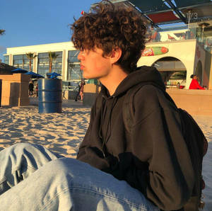 金色夕陽下，穿著黑色連帽衫坐在沙灘上的捲髮年輕人。