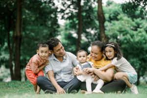一個微笑的五口之家坐在公園的草地上，周圍有樹木。 — 家庭 壁紙