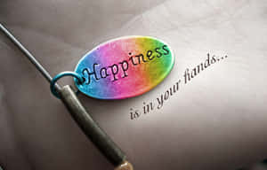 一個刻有「Happiness」的彩虹色飾物放在一隻手上，旁邊寫著「is in your hands...」。 — 喜悅 壁紙