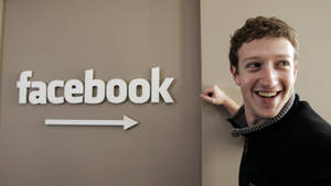 微笑的男人在米色牆上Facebook標誌和箭頭旁邊。 — 馬克·祖克柏 壁紙