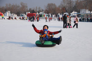 穿著紅色外套和頭盔的孩子在雪地上騎著綠色雪管，周圍有人。 — 火焰與冰 壁紙