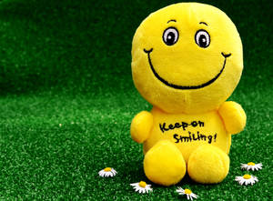 黃色毛絨笑臉玩具，帶有「Keep on Smiling!」文字，坐在草地上有雛菊。 — 正向 壁紙