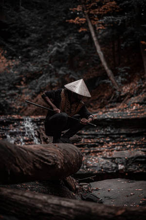 戴圓錐形帽子的人蹲在森林溪流附近的圓木上，手持長杖。 — 混沌武士 壁紙