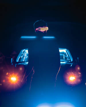 戴著發光眼鏡的人站在一輛車頭燈明亮的汽車前，夜晚。 — 深色霓虹 iPhone 壁紙