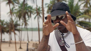 戴太陽眼鏡和漁夫帽的男人在沙灘上做手勢，背景有棕櫚樹。 — Skepta 壁紙