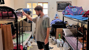 男子在雜亂的宿舍房間裡整理衣服，房間裡有雙層床。 — 臥室 壁紙