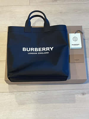 黑色 Burberry 手提袋帶有標誌，放在木地板上，旁邊是品牌盒子。 — Burberry 壁紙